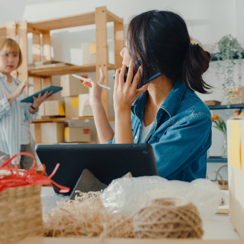 Young Asia businesswomen using mobile phone call receiving purchase order and check product on stock work at home office. Small business owner, online market delivery, lifestyle freelance concept.